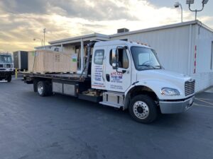 An Affordable Towing LLC flatbed tow truck transporting a large wooden crate at dusk in Anaheim, CA.