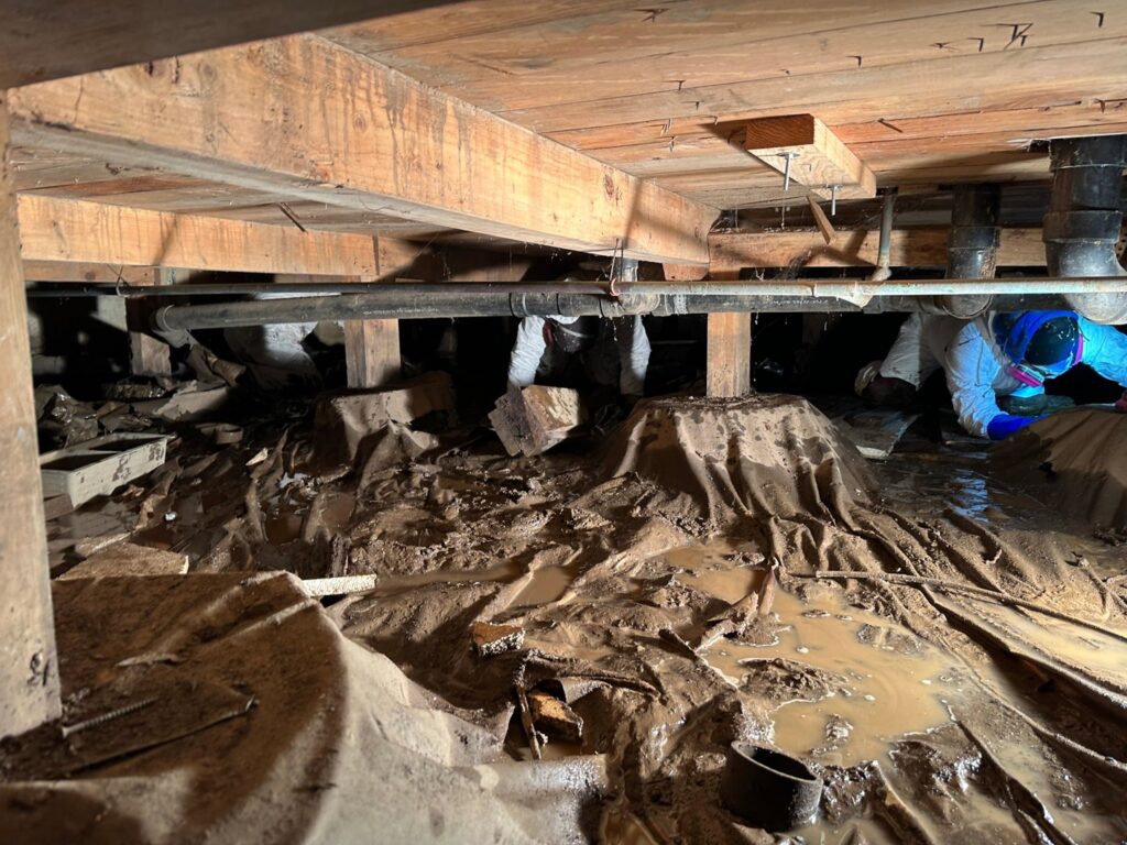 Workers in protective gear performing water damage remediation in a flooded and muddy crawl space for The Recover Tech Restoration in Vancouver, WA.