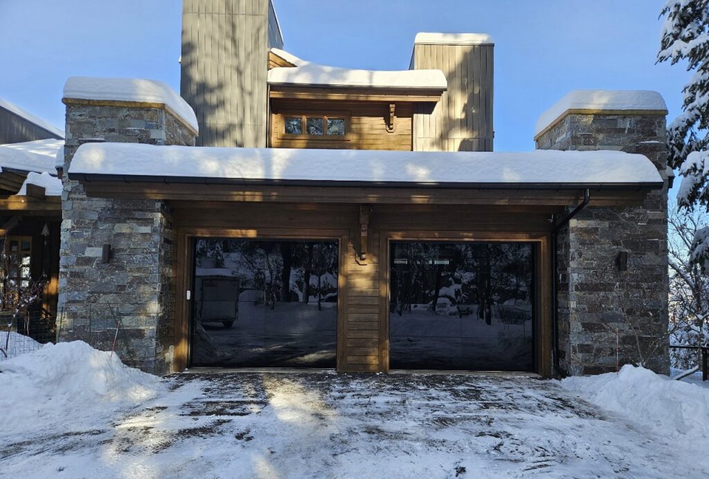 Custom garage doors on a stone and wood house in a winter setting, installed by Riverside Garage Doors in Kalispell, MT.