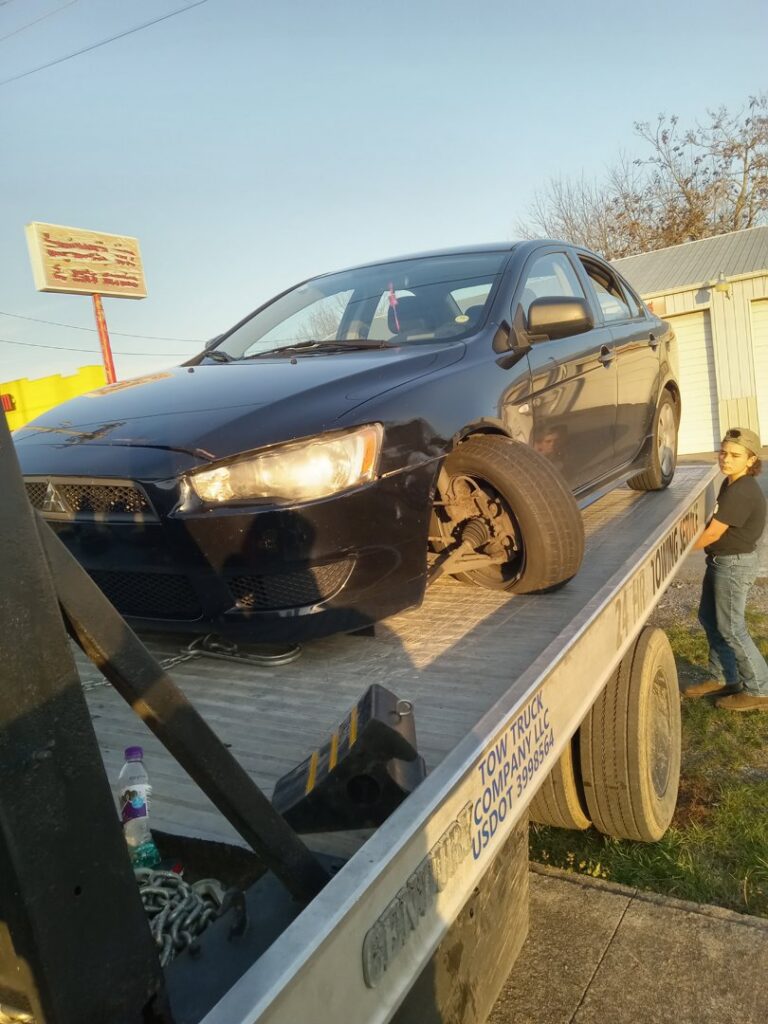 A damaged black car with a broken wheel loaded onto a flatbed tow truck by Tow Truck Company in Johnson City, TN.