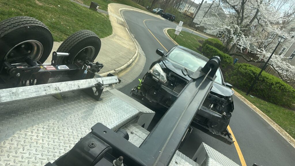 A heavily damaged black car positioned on the bed of a tow truck, ready for transport by Ricky's Towing & Recovery LLC in Derwood, MD.