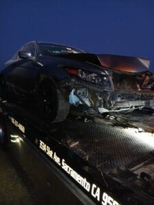 A damaged dark car being towed by an AJ's Towing Services & Roadside Assistance flatbed truck in Sacramento, CA.