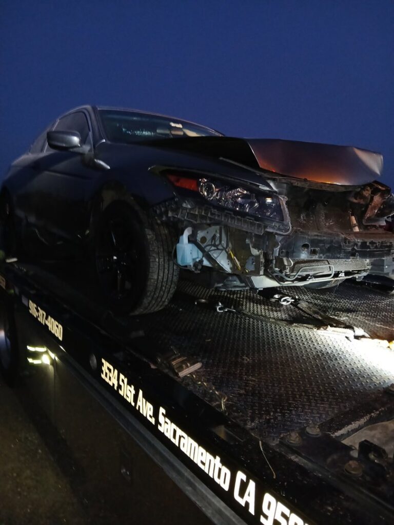 A damaged dark car being towed by an AJ's Towing Services & Roadside Assistance flatbed truck in Sacramento, CA.