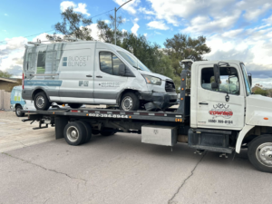 A damaged Budget Blinds commercial van being transported on a flatbed tow truck by Trejo's Towing LLC in Phoenix, AZ.