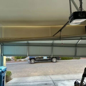A damaged garage door partially open, with a work truck visible outside, indicating a repair job by Rebel Overhead Doors in Las Vegas, NV.
