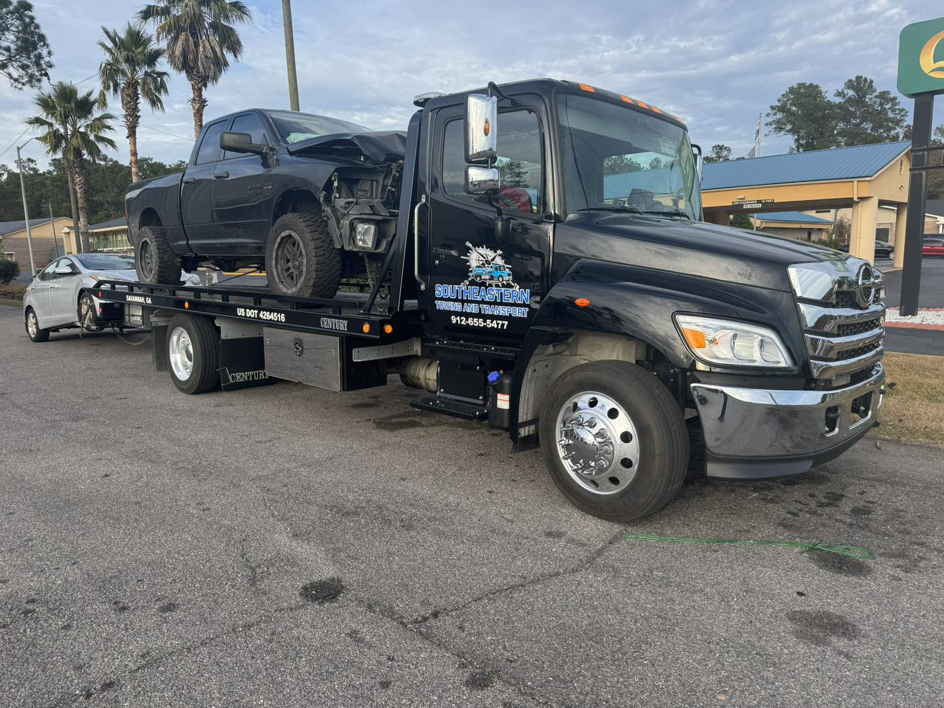 A black tow truck from Southeastern Towing and Transport towing a damaged pickup truck and another car in Savannah, GA.