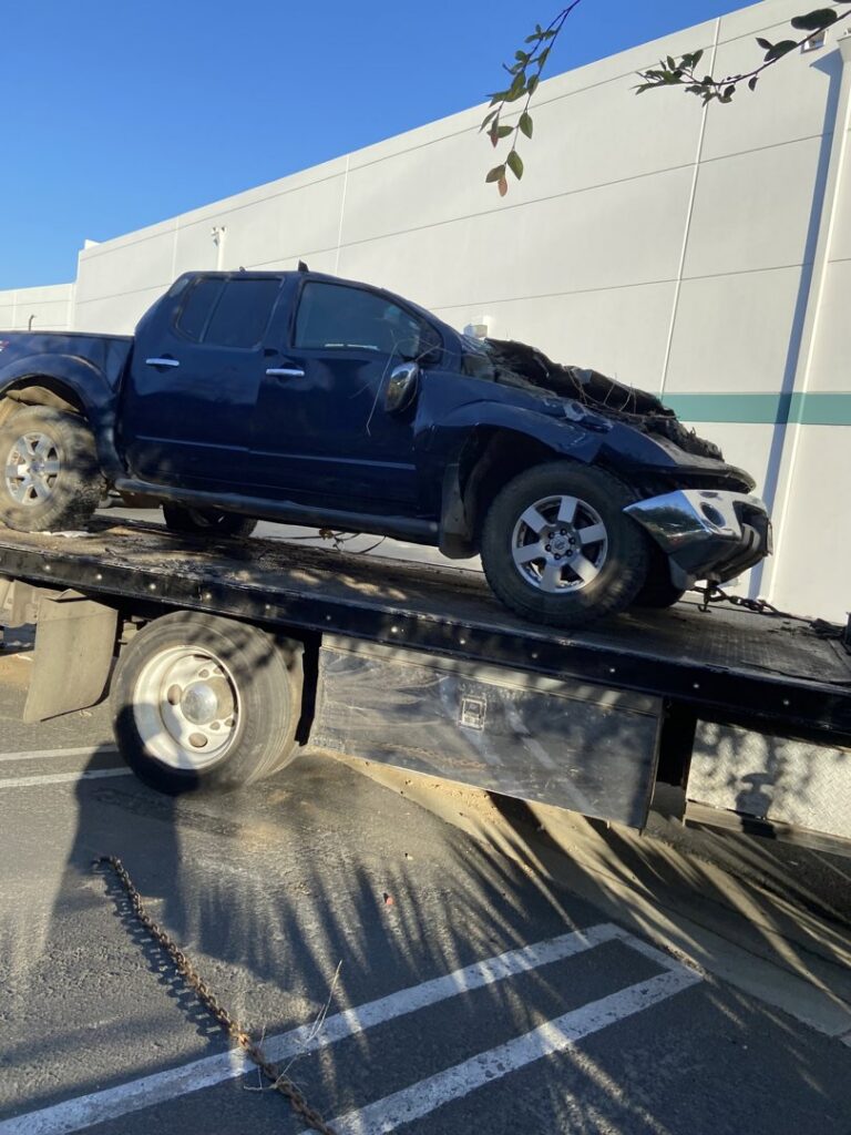 An Affordable Towing LLC flatbed tow truck transporting a damaged dark blue pickup truck in Anaheim, CA.