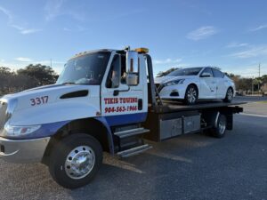 A damaged white sedan being transported on a flatbed tow truck by Tiki's Towing LLC in Jacksonville, FL.
