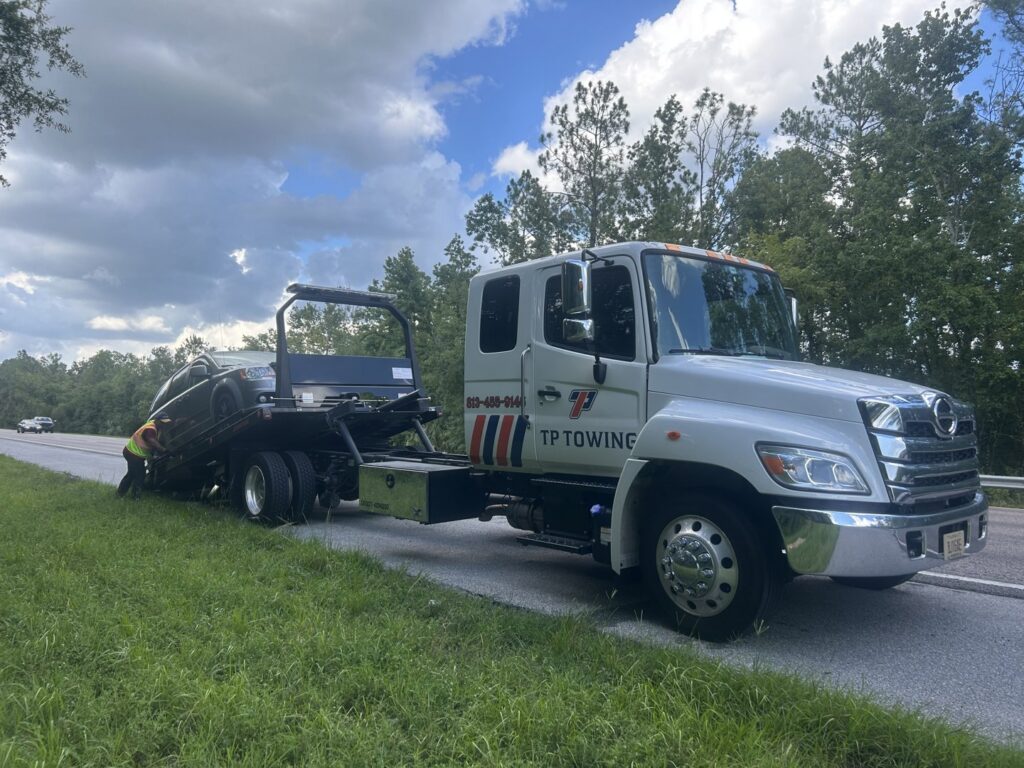 A dark car being loaded onto a TP Towing flatbed truck from the roadside, offering recovery services in Tampa, FL.