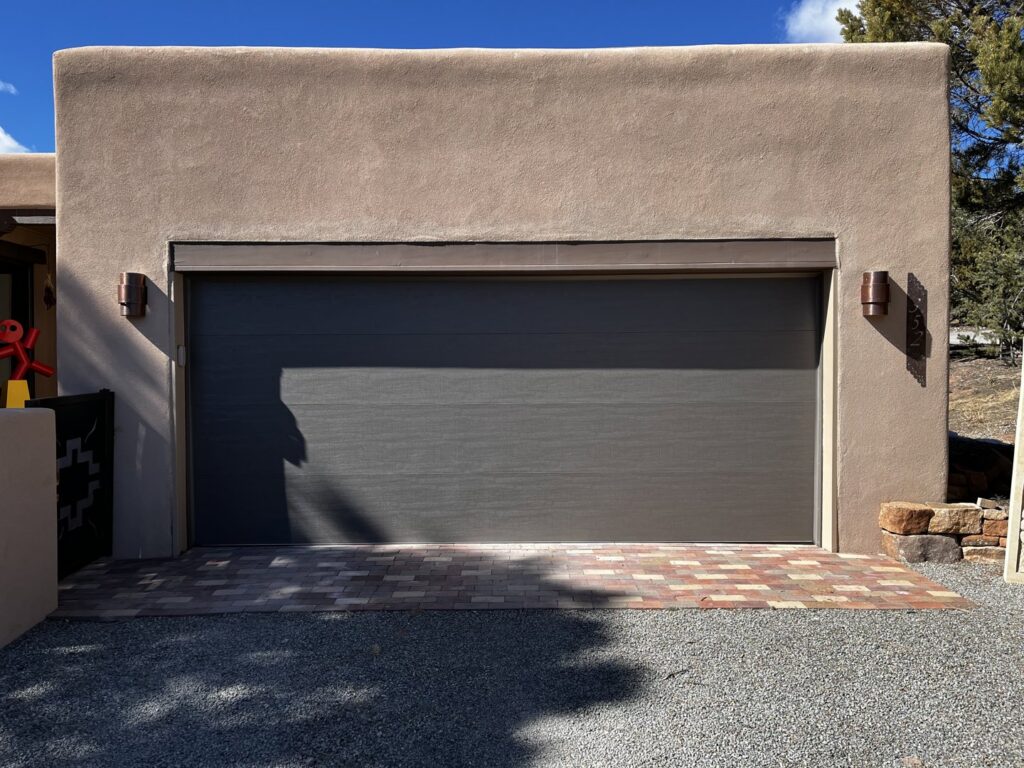 A dark gray garage door installed on a stucco home with a brick paver driveway by Precision Garage Door of Albuquerque and Santa Fe, NM.