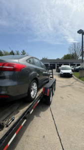 A dark grey car being towed on a flatbed trailer in front of an auto shop by 82Hauling & More LLC in St. Louis, MO