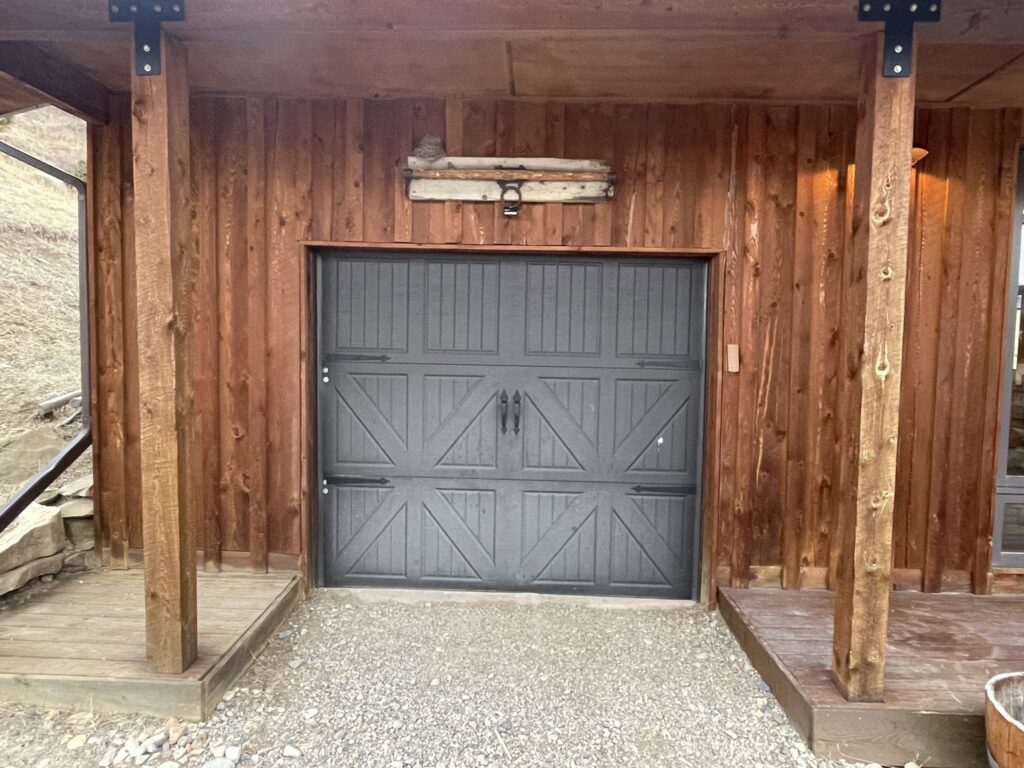 A dark grey panel garage door with decorative hardware installed on a rustic wooden building by Elite Garage Door Service & Repair in Billings, MT.
