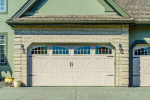 A decorative beige garage door with windows and hardware on a brick house by PS Garage Doors of Williston, ND.