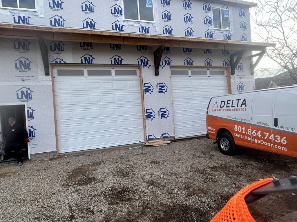 A Delta Garage Door service van parked at a new construction site with two white garage doors installed in Millcreek, UT.
