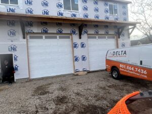 A Delta Garage Door service van parked at a new construction site with two white garage doors installed in Millcreek, UT.
