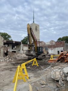 A demolition site with an excavator from DeCarlo Demolition Company and a church steeple in the background in Des Moines, IA.
