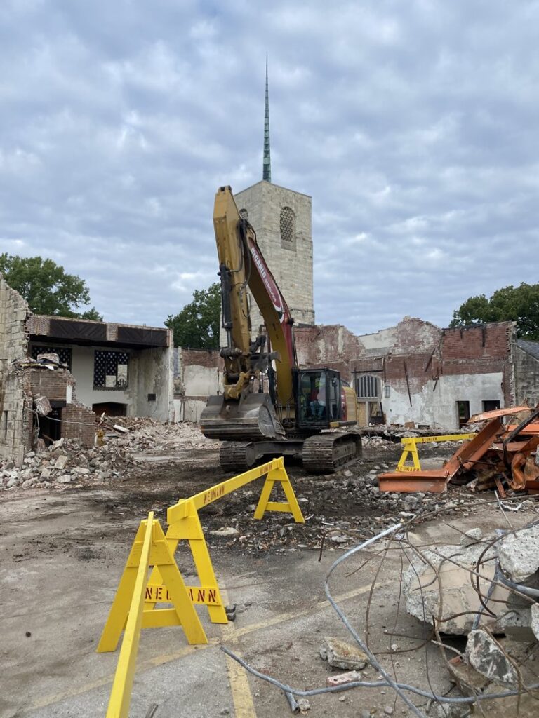 A demolition site with an excavator from DeCarlo Demolition Company and a church steeple in the background in Des Moines, IA.