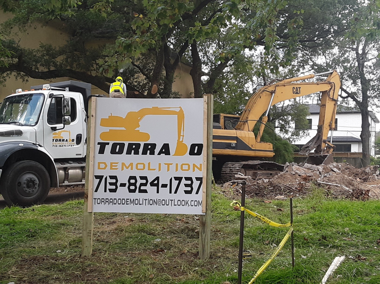 A demolition site with an excavator, dump truck, and Torrado Demolition business sign in Houston, TX.