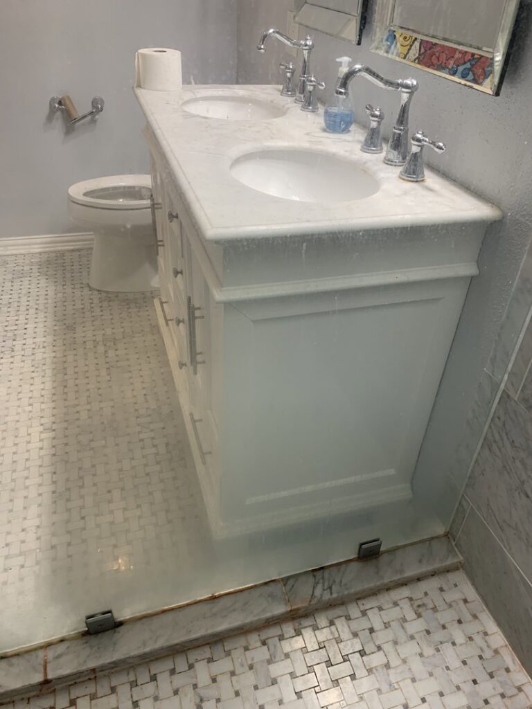 Dirty bathroom vanity with two sinks and glass shower door showing water spots, for cleaning by Jolley Services, INC in Bossier City, LA.