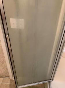 A cloudy glass shower door covered in water spots and soap scum, before cleaning by Quality Cleaning in Summerville, SC.