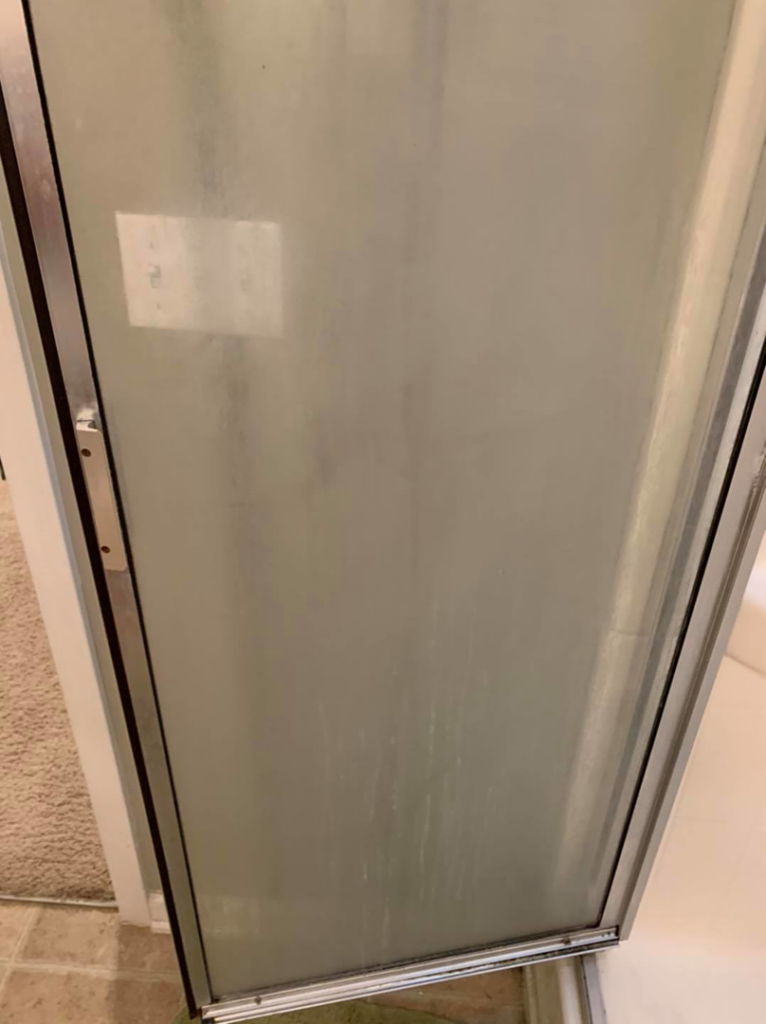 A cloudy glass shower door covered in water spots and soap scum, before cleaning by Quality Cleaning in Summerville, SC.