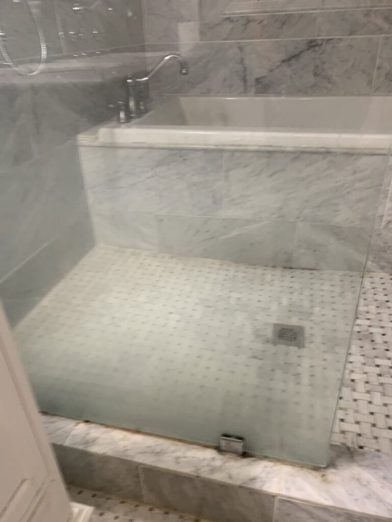 A glass shower enclosure with visible water spots and residue, indicating a cleaning job by Jolley Services, INC in Bossier City, LA.