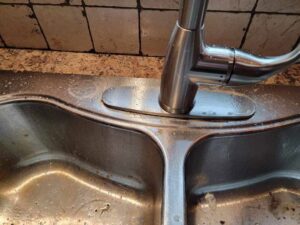 A dirty stainless steel kitchen sink and faucet with water spots and grime, showing a 'before' cleaning state by Sparkle & Shine of Iowa in Urbandale, IA.