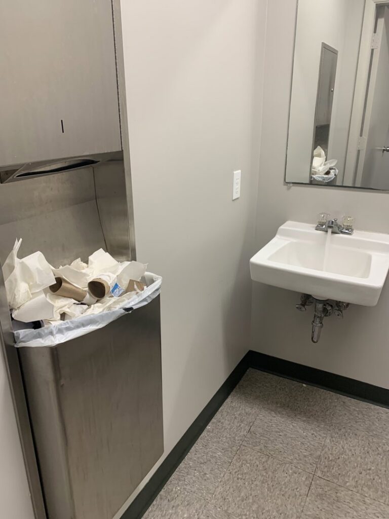 A dirty public restroom with an overflowing trash can and messy sink, before cleaning by West & Co Cleaning Services in Laurel, MD