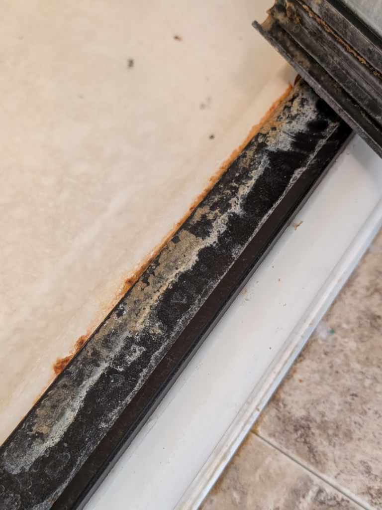 A close-up of a heavily soiled shower door track with grime and mold buildup, showing a 'before' cleaning state by White Rose Cleaning in Idaho Falls, ID.
