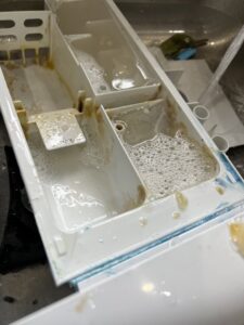 A dirty washing machine detergent dispenser filled with soapy water and grime, indicating a deep cleaning task by Little Cubs Cleaning Services in Rochester, MN.