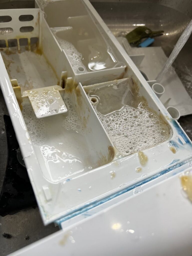 A dirty washing machine detergent dispenser filled with soapy water and grime, indicating a deep cleaning task by Little Cubs Cleaning Services in Rochester, MN.