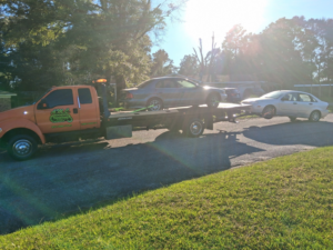 An orange AMC Towing flatbed truck in Mobile, AL, transporting one sedan and towing another behind it.