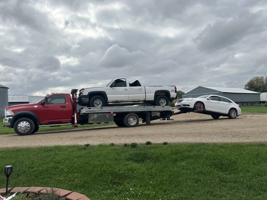 A red tow truck performing a double tow, carrying a pickup and towing a sedan, for Geely Towing in Fargo, ND.