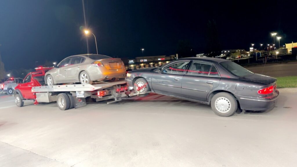 A red tow truck performing a double tow, carrying one sedan and towing another, for Geely Towing in Fargo, ND.