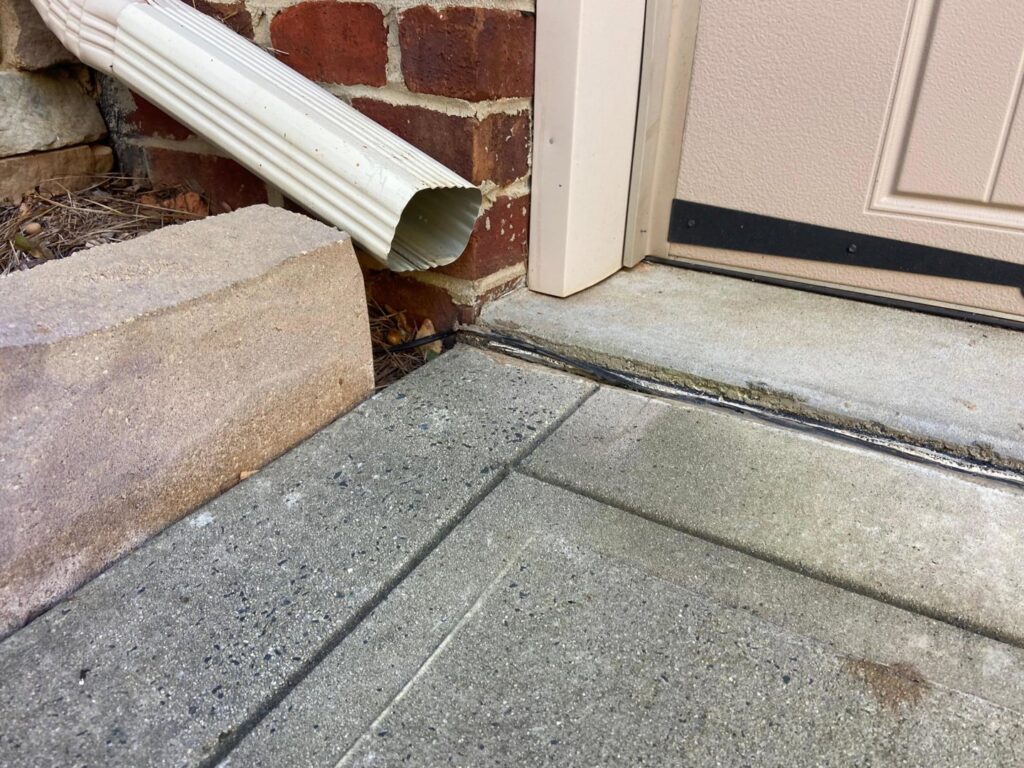 A downspout discharging water onto a concrete path, highlighting a potential drainage issue for HNO Waterproofing & Slab Lifting Pros in Concord, NC.