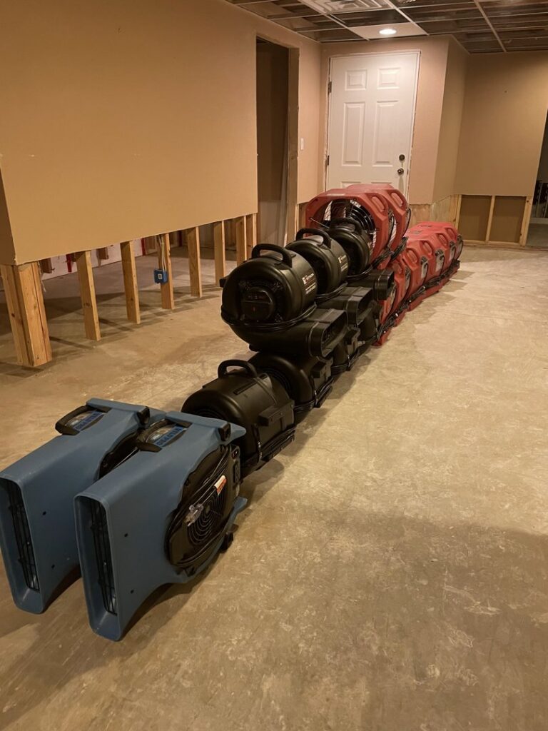 A row of industrial drying equipment used for water damage restoration in a room with stripped walls, provided by The Restoration Experts in Waupaca, WI.