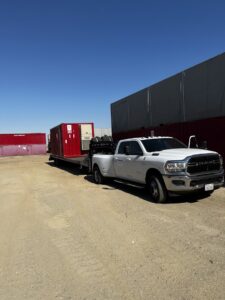 A white dually pickup truck towing a flatbed trailer loaded with large red containers for Inglewood Transmission in Long Beach, CA.