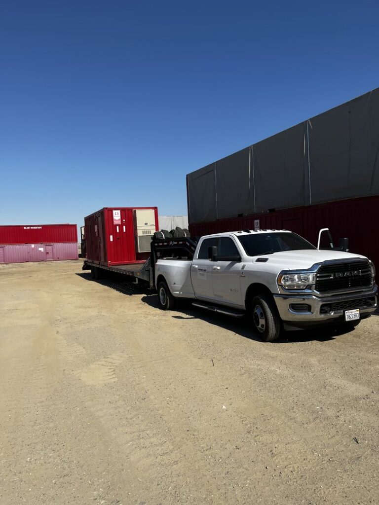 A white dually pickup truck towing a flatbed trailer loaded with large red containers for Inglewood Transmission in Long Beach, CA.
