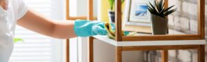 A hand in a blue glove wiping a wooden shelf with a yellow cloth, demonstrating dusting services by Orange Cleaning Services in Stamford, CT.