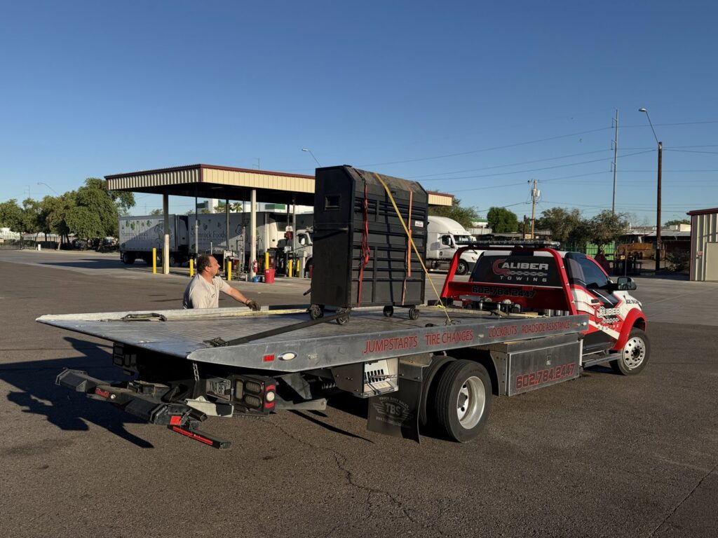 A large black equipment cabinet being transported on a flatbed tow truck by Caliber Towing in Phoenix, AZ.