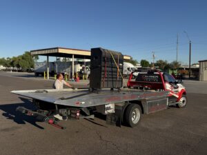A large black equipment cabinet being transported on a flatbed tow truck by Caliber Towing in Phoenix, AZ.