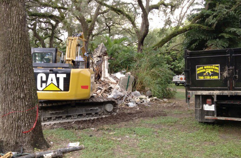 A CAT excavator demolishing a structure with a dump truck nearby for Dade Wrecking Demolition LLC in Miami, FL.