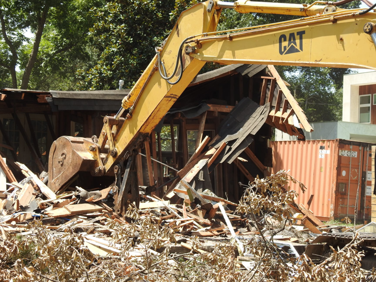 An excavator bucket actively demolishing a wooden house structure, performed by Torrado Demolition in Houston, TX.