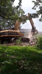 An excavator with a grapple attachment actively tearing down a structure, generating dust, by Torrado Demolition in Houston, TX.