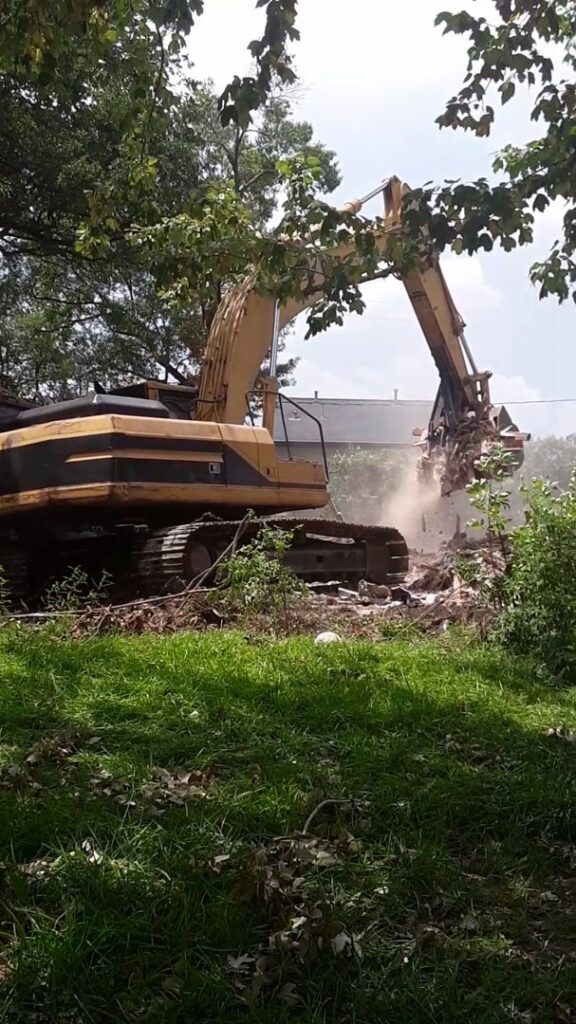 An excavator with a grapple attachment actively tearing down a structure, generating dust, by Torrado Demolition in Houston, TX.