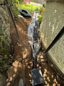 Installation of exterior drainage pipes and a grate in a trench next to a building foundation by Peach3 Group in New Rochelle, NY.
