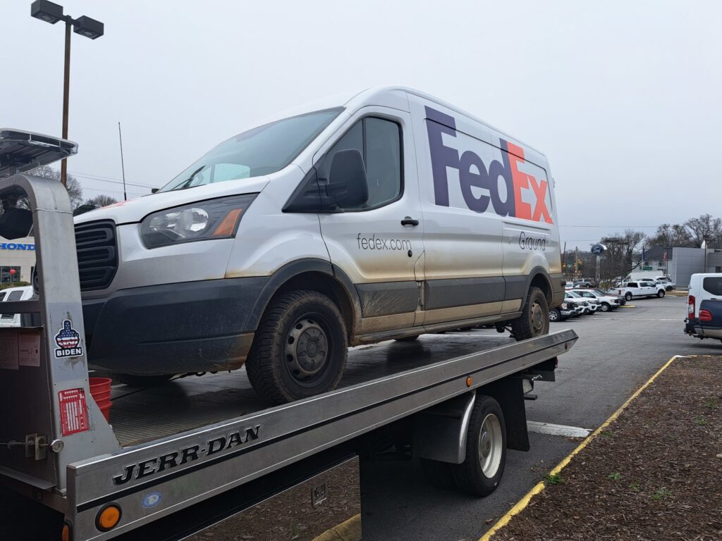 A white FedEx van being transported on a flatbed tow truck by Acme Towing Services in Greenville, SC.