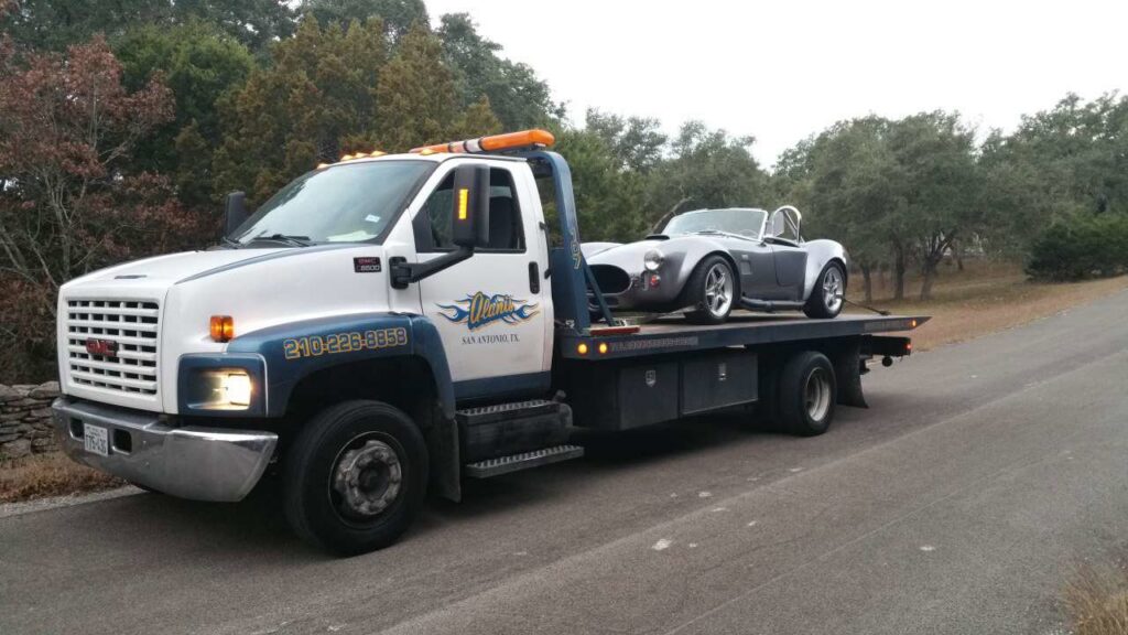 A flatbed tow truck from Alanis Wrecker Service carrying a grey sports car in San Antonio, TX.