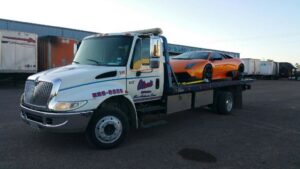 A flatbed tow truck from Alanis Wrecker Service carrying an orange sports car in San Antonio, TX.