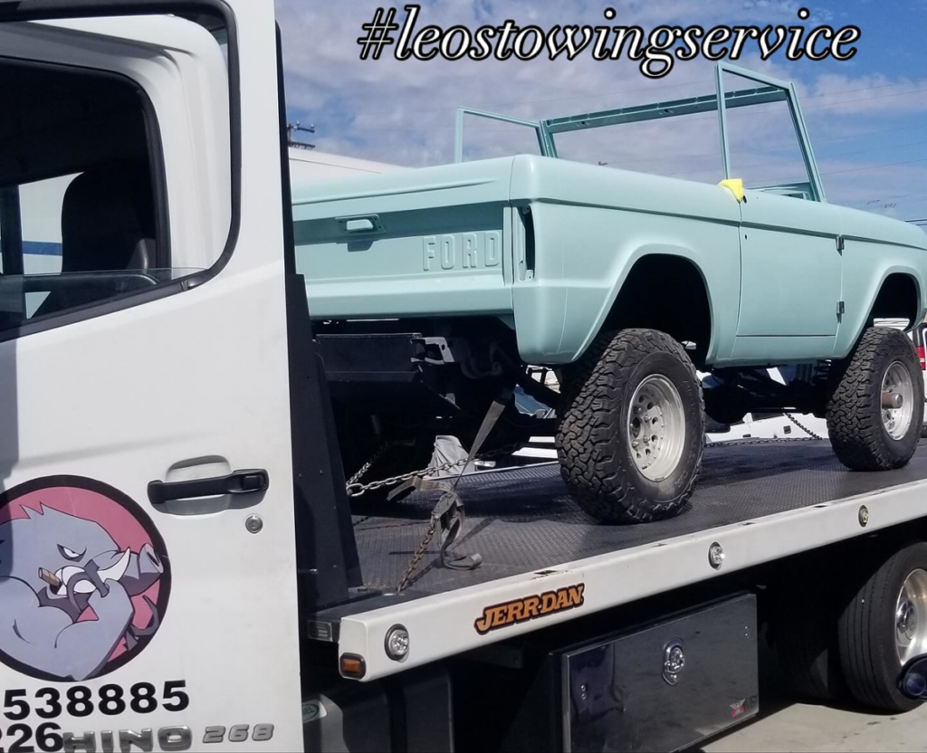 A flatbed tow truck from Leos Towing Service carrying a vintage light blue Ford Bronco body in Anaheim, CA.